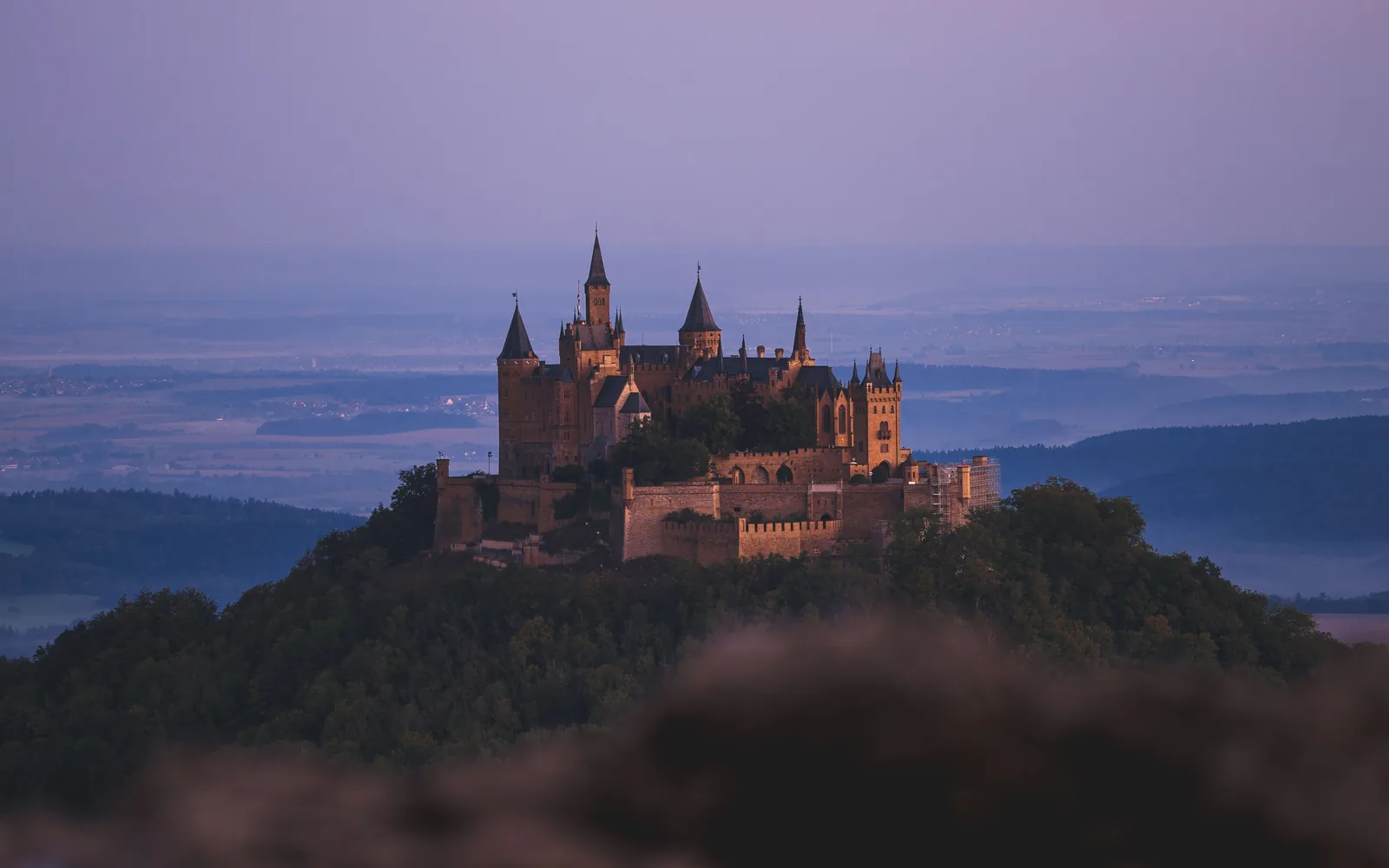 Il Burg Hohenzollern sulla sua sommità boscosa al crepuscolo, le torri illuminate sopra la pianura sveva